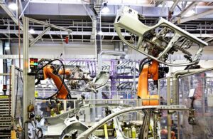 Two orange industrial robot arms lifting large car parts in a mostly white factory.