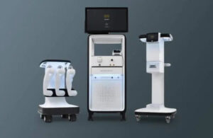 From left to right, a white platform on wheels with three robotic arms, a monitor on a white stand and another white and black stand.