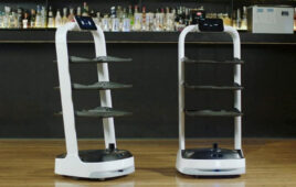 Two white serving robots with three black shelves and a screen with eyes.
