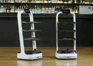 Two white serving robots with three black shelves and a screen with eyes.