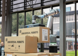 A Universal Robots cobot with a Robotiq gripper lifting a box.